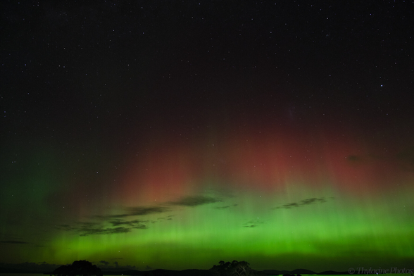 An aurora fills most of the lower half of the frame, with a red/pink glow above the typical green, closer to the horizon.  Distant water reflects the green sky, and tree lined hills are just visible in the distance.  The cloud has thinned out almost completely compared to the previous images, a few sparse patches to the left.