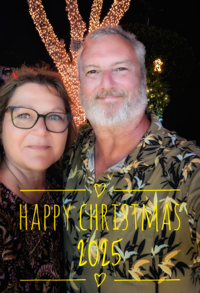A festive selfie of a man with a grey beard wearing a green patterned shirt and a woman with glasses. They are smiling in front of a tree wrapped in bright golden Christmas lights at night. Overlaid yellow text reads "Happy Christmas 2025" accented with small hand-drawn hearts and horizontal lines.