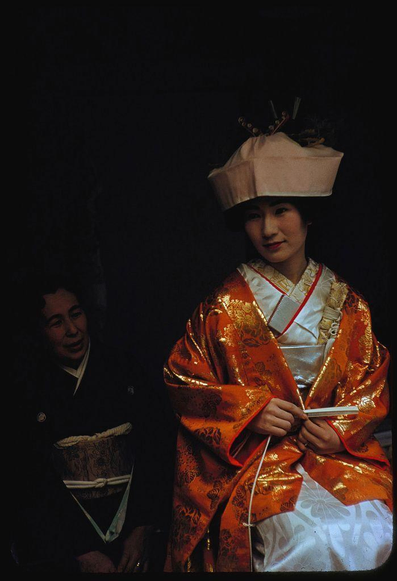 A person wearing a traditional Japanese kimono and headpiece is standing beside another individual in dark clothing. The main subject holds some thin objects, possibly chopsticks or decorative items, with both hands while looking directly at the camera with a slight smile. They are positioned against a black background that emphasizes their vibrant orange garment adorned with golden patterns.