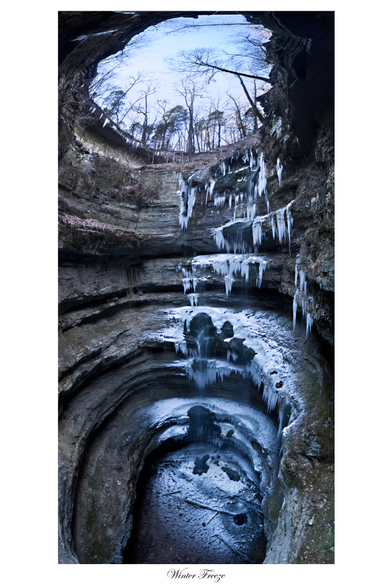 White framed image titled "Winter Freeze": a vertical panoramic created from hanging on rope in a deep entrance pit to a cave in the middle of winter so the waterfall on the far side is frozen into ice stalagmites and snow blankets the ledges around the pit and bottom. 