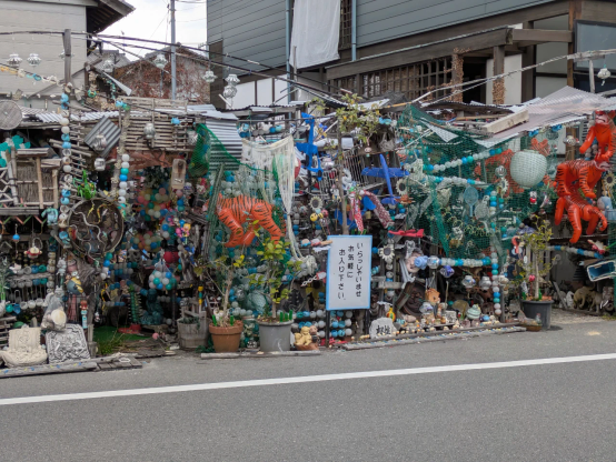 住宅地の中にあるガラクタで埋もれた空間。「いらっしゃいませ　お気軽にお入り下さい」という看板が立っている。広島県竹原市。