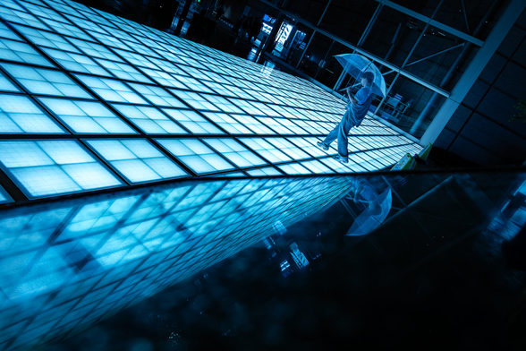 Reflection shot of a man with umbrella walking on a lit glass esplanade in Tokyo at night. The chosen angle leaves the impression that the man could fall of a flying structure. The scene is bathed in dark turquoise tones and surrounded by the night, in a cyberpunk atmosphere.