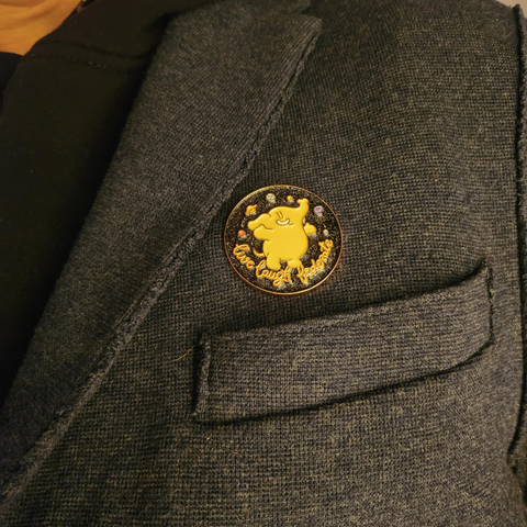 Rund Mastodon-pin festet på jakkeslag. Pinnen viser den gule Mastodon-figuren som danser foran en stjernehimmel med små planeter, og teksten “Live Laugh Federate” nederst. Bakgrunnen er mørk, og pinnen er plassert på en mørkeblå ulljakke.