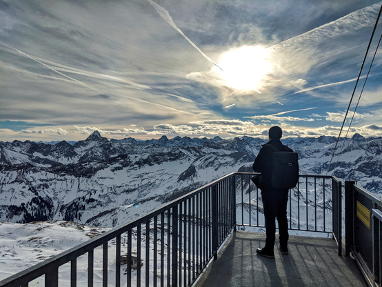 Panoramic view of the alps on top of the Nebelhorn in Germany. Flat clouds with a large hole with the sun seeping through seen right above me who is standing slightly to the right in the picture. You can see the ropes of the gondola on the right and I’m looking out to take in the beautiful view. 