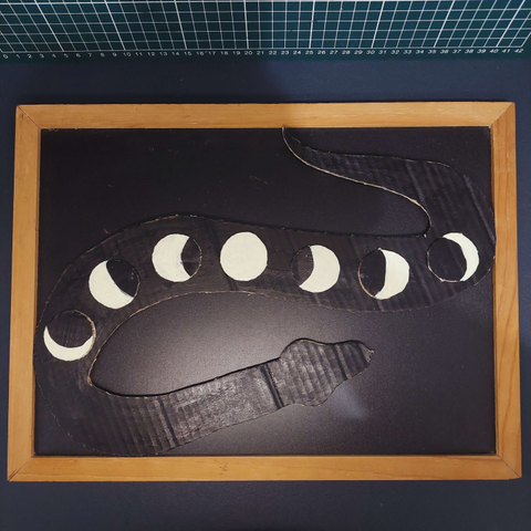 A cardboard cutout of a snake in silhouette, with the phases of the moon along its body. The snake is painted black and displayed upon a black background so you only see its shape in texture, highlight and shadow.