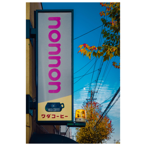A color photograph of a tall vertical café sign mounted on the side of a building. The sign reads “nonnon” in large pink letters, with a small logo and Japanese text for Wada Coffee near the bottom. Beneath the sign is a yellow rotating warning light. In the background, power lines crisscross a bright blue sky, a pale daytime moon is visible, and autumn leaves in shades of yellow and red frame the right side of the image.