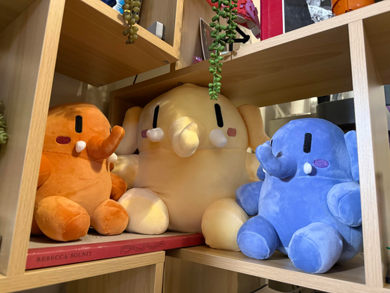 Three Plushtodon mastodons in peach, light yellow, and blueberry are sitting on my bookshelf. They look heroic and ready to face the new year. They are surrounded by potted plants and other tchotchkes.