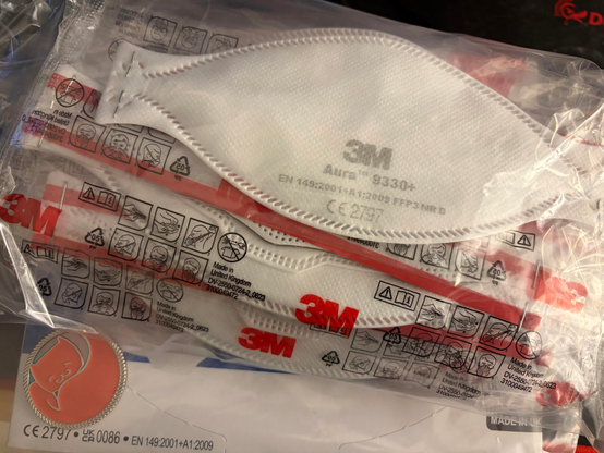 a stack of 10 3M Aura 9330+ face masks wrapped in plastic. in the bottom left corner is the .meow challenge coin from 39c3 