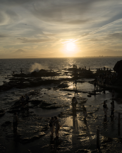 Photograph of a rocky shore, the sea and the sun setting, with the silhouette of Mount Fuji visible over the horizon. 
Small groups of people can be seen in backlight standing on the wet rocky shore, their long shadows projected by the setting sun. 
A wave is breaking on the end of the shore, creating a large splash.
This is a larger, uncropped, portrait-format version of the previous picture.