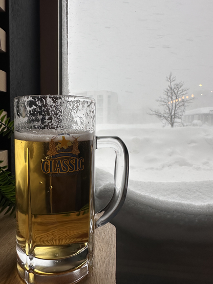 A pint of beer on a table with a window behind it showing snow dumping down.