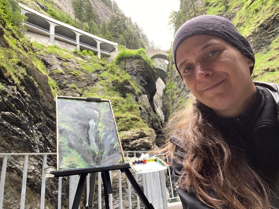 Die Künstlerin steht lächelnd mit schwarzer Kappe und langen Haaren neben ihrer Feldstaffelei tief unten in der Viamala Schlucht. Links oben ist die Hauptstrasse, in den Fels gebaut und mit einer Gallerie vor Steinschlag geschützt. Oben in der Mitte überqueren zwei Brücken die steile, felsige Schlucht. Auf der Staffelei steht das fertige Bild, welches auf realistische Weise die Viamalaschlucht abbildet. Die Felsen sind dunkelgrau und die spärliche Vegetation wie Moos, Gräser und kleine Büsche sind knackig grün.