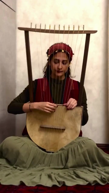 A lady in traditional dress: a long, olive green skirt, a black top, red waistcoat and headscarf, sitting on the floor playing a harp.