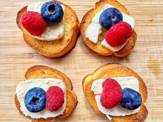 木製のカッティングボードの上に並べられた小さなトースト。カリッと焼いたバゲットに白い Brie チーズをのせ、ラズベリーとブルーベリーをトッピングした一口サイズのオープンサンドが複数並んでいる。

Small toasted baguette slices arranged on a wooden cutting board, each topped with creamy white Brie cheese and garnished with raspberries and blueberries, presented as bite-sized open sandwiches.