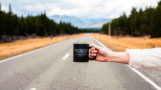 Hand holding a black coffee mug with text Dream Sleeper Mini and its logo on it. Arm is stretched while standing on the side of the road so the mug is "floating" above the road. New Zealand scenery in the background.