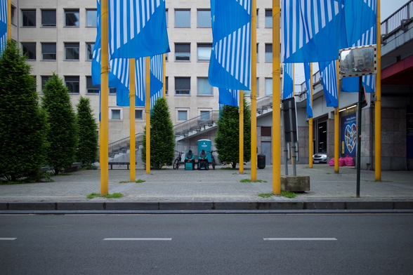 Two bicycle couriers waiting for orders in a plaza in Brussels, Belgium. The plaza is full of blue banners hung from yellow masts.