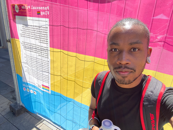 Selfie in front of pansexual pride flag on sunny day in Munich, Germany. Listed are the definitions of pansexuality in multiple languages (if you look really close, and reverse the text). :)