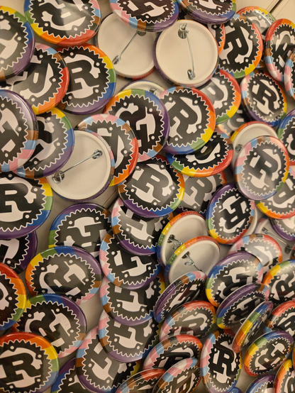A whole pile of pin buttons with the Rust logo on top of the progress pride flag.