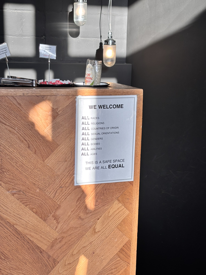 A photo of a white sign taped to the side of a wooden counter with a herringbone pattern in a cafe. Sunlight and shadows are cast across the sign and the wood. The sign reads in black uppercase text: “WE WELCOME ALL RACES, ALL RELIGIONS, ALL COUNTRIES OF ORIGIN, ALL SEXUAL ORIENTATIONS, ALL GENDERS, ALL BODIES, ALL ABILITIES, ALL AGES. THIS IS A SAFE SPACE. WE ARE ALL EQUAL.” In the background, hanging industrial-style lights and condiments on the counter are visible.