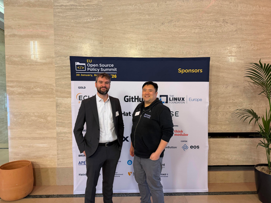 Paul Sharratt and Powen Shiah at the EU Open Source Policy Summit. They’re standing in front of a large wall with the summit logo and some sponsor logos are visible behind them.