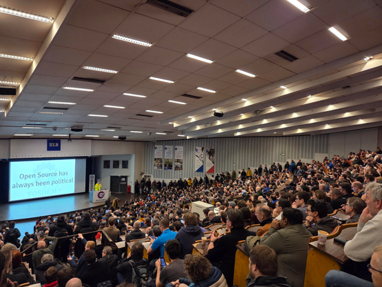 Large, packed auditorium with "Open Source has always been political" on the screen. 