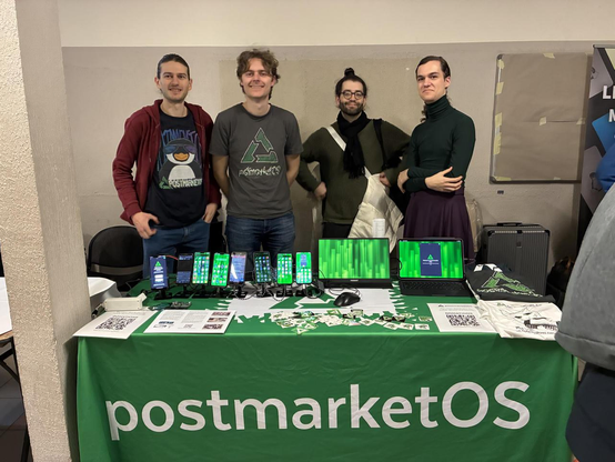 4 awesome postmarketOS team members behind our FOSDEM booth. The booth has a table cloth on it showing the project name and displays various phones, stickers, tablets and t-shirts.
The table cloth is green with white text
