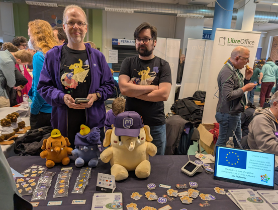 The mastodon table, with plushies, stickers, pins for sale and behind the table two of our people holding the fort.