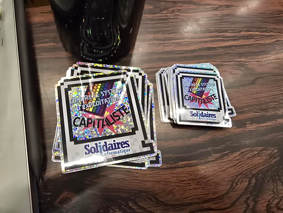 deux piles d'autocollants Solidaires Informatique, paillettes et arc en ciel, contre le système d'exploitation capitaliste