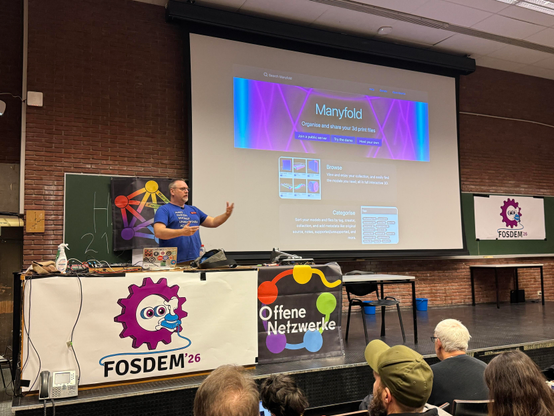 A presenter is speaking at a conference, standing in front of a screen displaying the "Manyfold" website, which organizes and shares 3D print files. Various logos and banners are visible, including "FOSDEM 26"