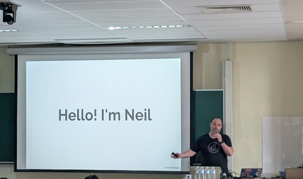 Neil, white man in a black t-shirt, standing at a lectern gesticulating at a slide which says "Hello! I'm Neil" 