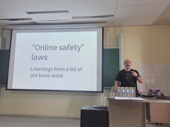 Neil on stage with the main slide next to him stating "Online Safety Laws, learnings from a lot of bro bono work"