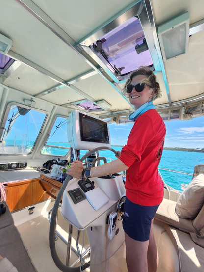 A curly haired woman wearing sunglasses and a red shirt is steering a sailboat with hands on the helm. The cockpit of the boat is visible, featuring a navigation screen and various controls. Clear blue water can be seen through the windows.