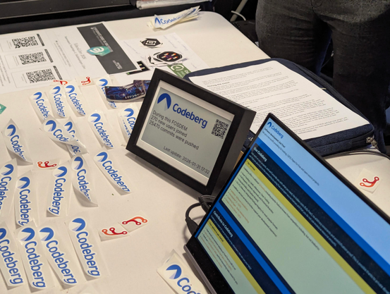 e-Paper depicting Codeberg's stats "since the beginning of FOSDEM".

During this FOSDEM, 270 new users joined. 25740 commits were pushed.

There's a display depicting stand.codeberg.org to the right, and many visible Forgejo and Codeberg stickers to the left.