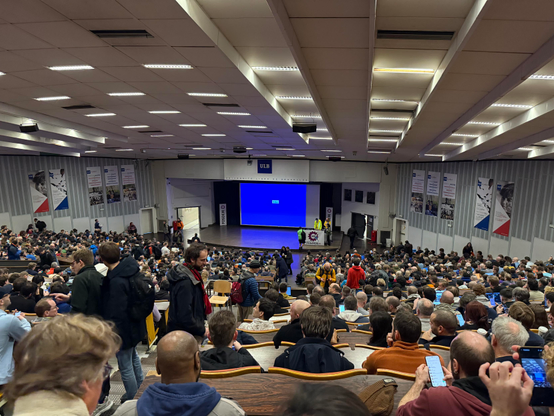 Photo from way up in the back of the Janson room showing a sea of back of heads, looking towards an empty stage. 