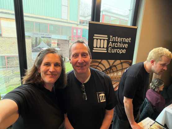 A man and a woman pose for a selfie as another man interacts with the public at the Internet Archive Europe stand at FOSDEM 