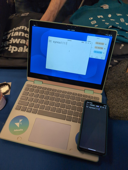 Laptop and phone with GNOME at the FOSDEM GNOME booth, both running reflection with a synced document.