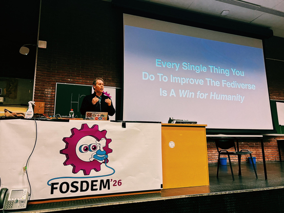 A photo showing Ben Pate on stage at FOSDEM in the Social Web devroom. He’s standing behind a table that has the FOSDEM ‘26 logo and next to him there is a large screen showcasing a slide that reads “Every Single Thing You Do To Improve the Fediverse Is A Win for Humanity”