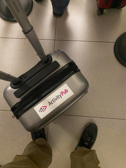 Looking down at my suitcase with a sticker with the ActivityPub logo on top, floor and shoes visible. Taken in an airport 