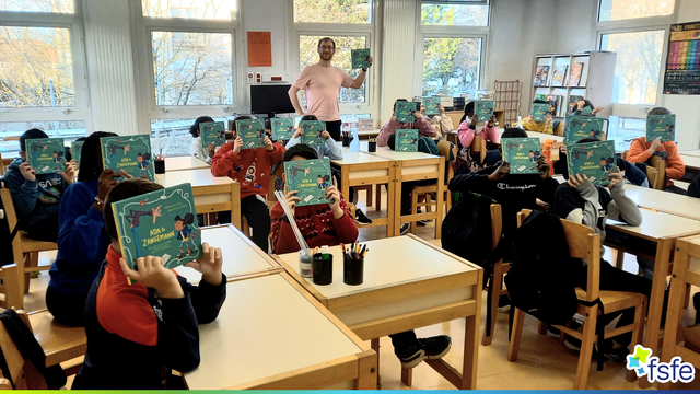 Group of children in a classroom with books in front of their faces + a teacher standing also holding the same book.