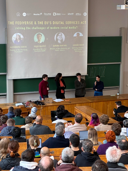A panel discussion is taking place in a lecture hall, featuring four speakers discussing "The Fediverse & the EU's Digital Services Act." The audience is seated, facing the speakers, while a presentation is displayed on a screen behind them: Alexandra geese, Felix hlatky, Sandra barthel, Jordan Maris. 