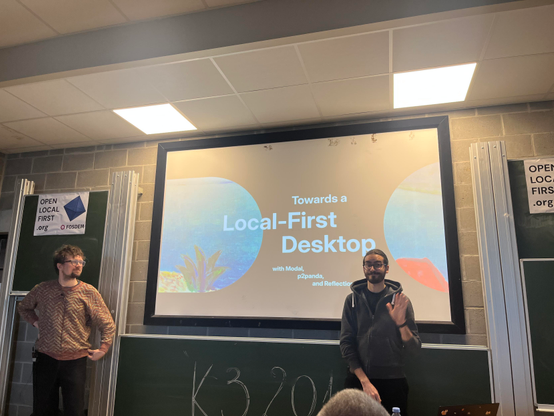 Photot: Two People standing infront of a presentation: "Towards a Local-First Desktop"