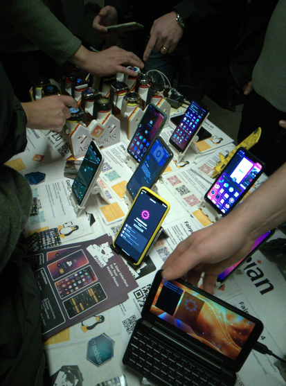 The image shows a table from the "Linux on Mobile" both at this year's FOSDEM.
Featured in the image are a variety of phones, smartwatches, flyers and stickers.

Also visible are the hands of people as they interact with the devices.