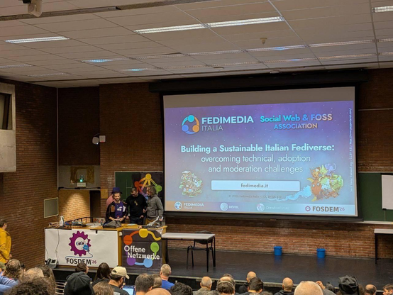 Un palcoscenico del FOSDEM ospita un ME, Francesco e Michele  dietro una scrivania, contro pareti in mattoni e luci soffuse a fluorescente. Uno schermo grande mostra “FEDIMEDIA ITALIA” e “Social Web & FOSS ASSOCIATION” in alto, seguiti dal titolo “Costruire un’Italia Sostenibile con il Fediverso: superando sfide tecniche, adozione e moderazione,” l’URL “fedimedia.it”, e loghi che includono “FOSDEM 26” e “Offene Netzerke”. Sul palco, banner mostrano “FOSDEM 26” e “Offene Netzerke”, mentre un pubblico si trova in primo piano. La stanza include un proiettore, ulteriori tavoli e secchi blu sul pavimento dello stage.
