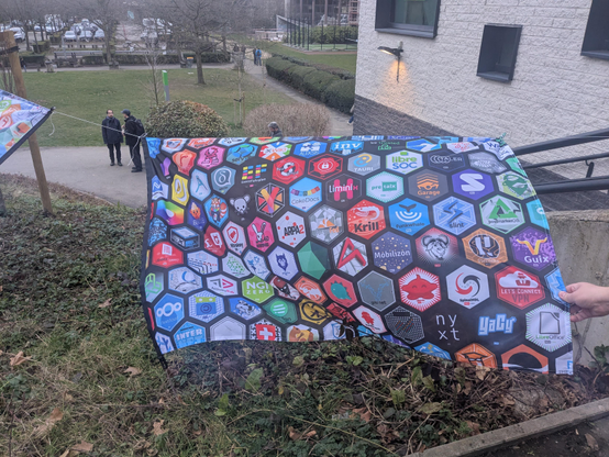 A photo of the NLNet hex stickers as a flag blowing in the wind with green grass in the background. The flag is being held by ChanHaeng Lee and Hong Minhee is pointing at Lemmy