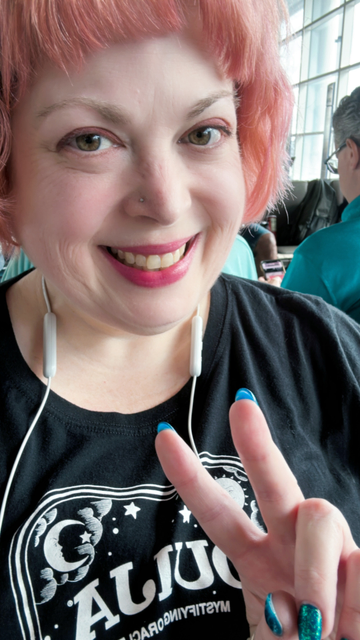A pink haired woman is taking a selfie and raising her fingers in a peace sign.