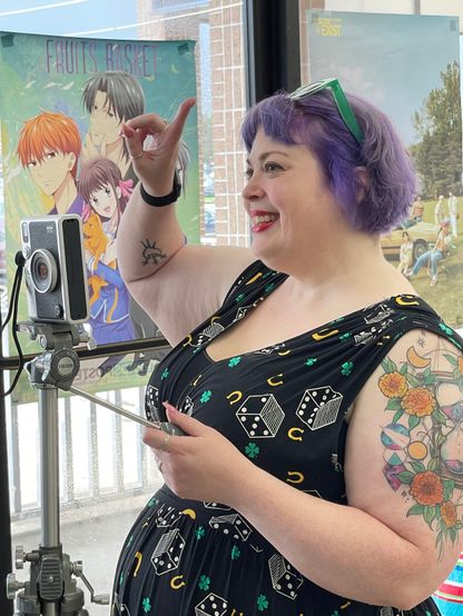 A purple haired woman stands behind a camera on a tripod, she is raising a finger and looking at someone to the left side.