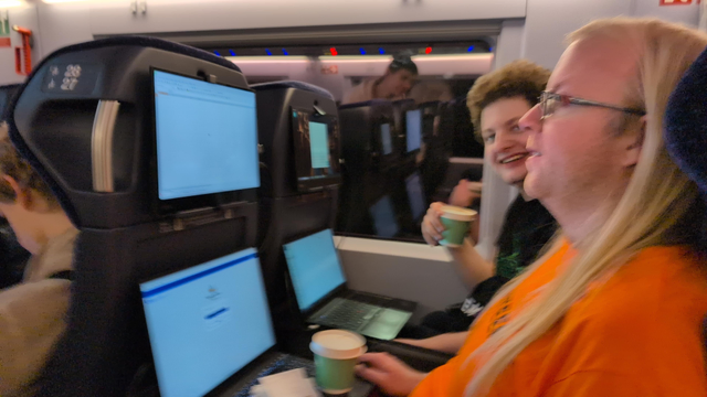 Two people on a train, each with a laptop on the table in front of them and an additional display mounted to the tablet holder in eyes' height.