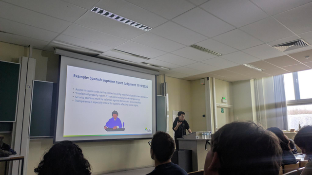 A person standing at a stage giving a talk with some slides an a screen behind him