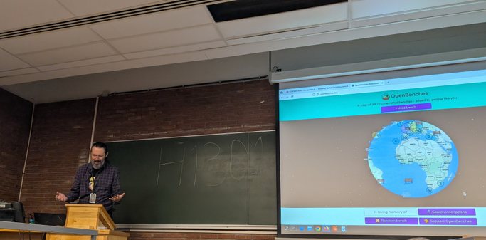 Terence Eden standing to the side of a projection of the Open Benches website 