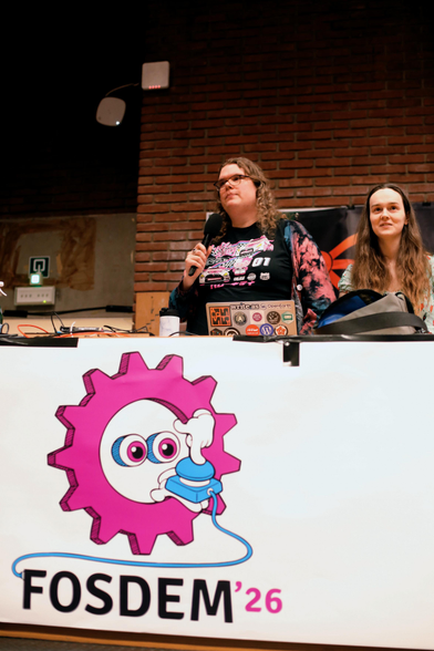 A photo showing Christine Lemmer-Webber and Jessica Tallon on stage at FOSDEM behind the podium with the logo "FOSDEM '26"