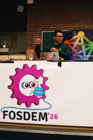 a vertical photo of Maho Pacheco on stage at FOSDEM. He's looking looking to the side while gesturing